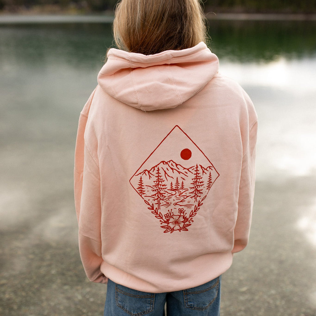 Person wearing a pink hoodie with a nature graphic, standing by a lake.