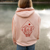 Person wearing a pink hoodie with a nature graphic, standing by a lake.