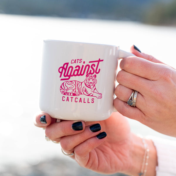 Person holding a mug with 'Cats Against Catcalls' design by a lake.